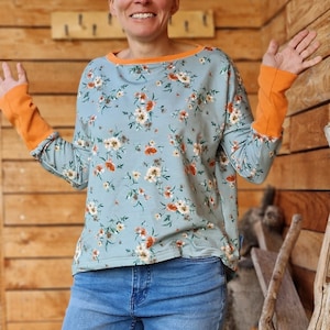 Könnte beinhalten: Ein langärmeliges, hellblaues Hemd mit Blumenmuster und orangefarbenem Besatz an Ausschnitt und Ärmelbündchen. Das Hemd wird von einer Person mit Brille getragen und zu einer blauen Jeans kombiniert.