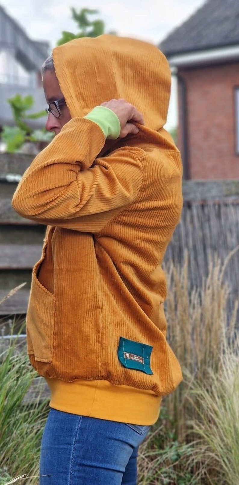 May include: A mustard yellow corduroy hooded sweatshirt featuring a large front pocket, a lime green cuff on the sleeve, and a small rectangular teal fabric patch with white text stitched onto the lower side of the garment.