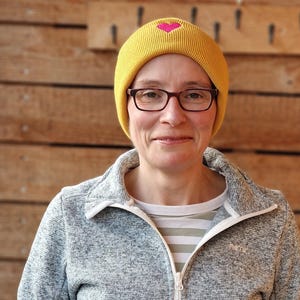 May include: A woman wearing a yellow beanie with a pink heart design, eyeglasses, and a gray zip-up jacket. The background is a wooden wall.