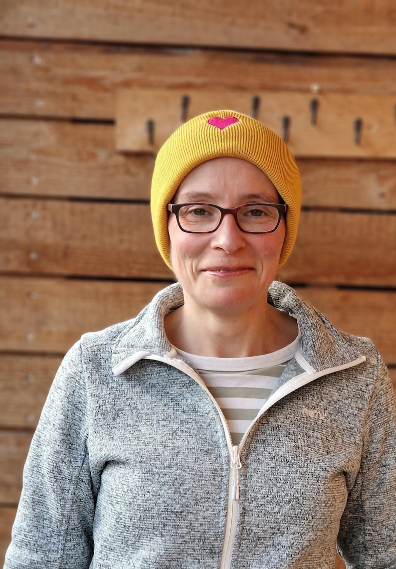 May include: A woman wearing a yellow beanie with a pink heart design, eyeglasses, and a gray zip-up jacket. The background is a wooden wall.
