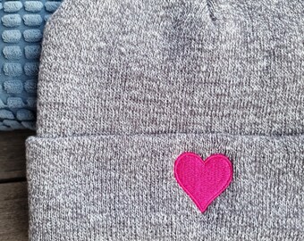 May include: A gray knit beanie with a bright pink heart embroidered on the front. The hat is folded at the brim and appears to be made of a soft, textured material. The background is a light blue textured surface.