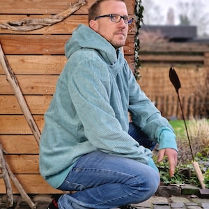 Op de afbeelding: Een persoon draagt een lichtblauwe corduroy hoodie met een capuchon, blauwe jeans en zwarte sneakers. De persoon zit op een bakstenen oppervlak en kijkt naar rechts.
