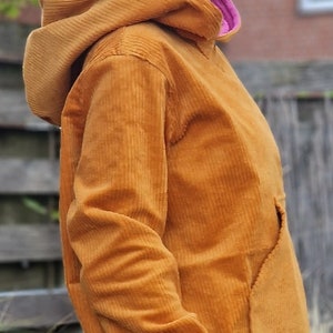 May include: A mustard yellow corduroy hooded sweatshirt featuring a large front kangaroo pocket, long sleeves with light green ribbed cuffs, and a bright pink inner lining visible at the neckline, worn over blue denim jeans.
