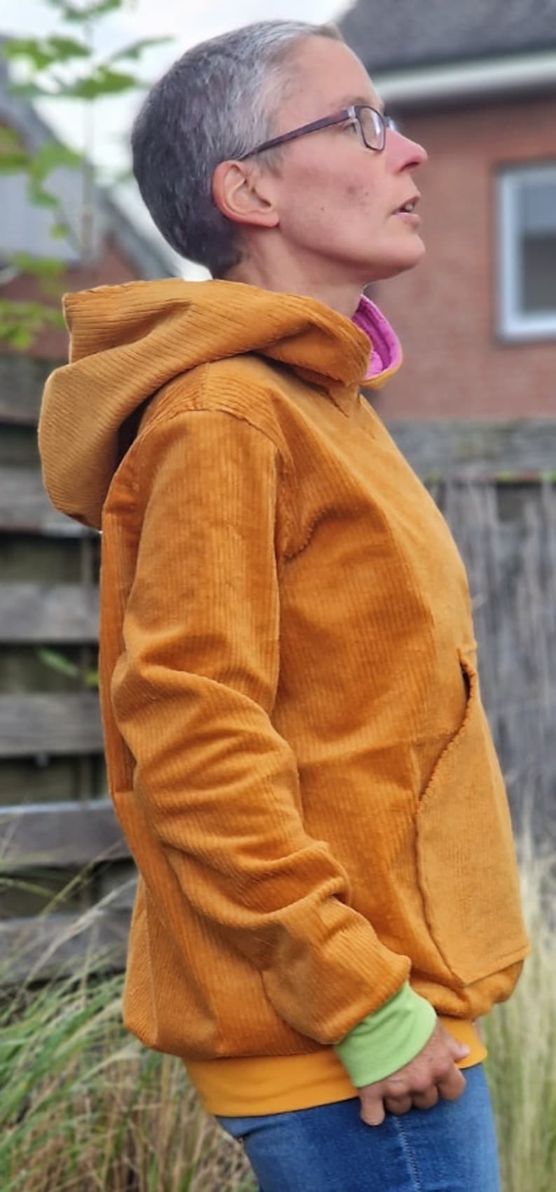 May include: A mustard yellow corduroy hooded sweatshirt featuring a large front kangaroo pocket, long sleeves with light green ribbed cuffs, and a bright pink inner lining visible at the neckline, worn over blue denim jeans.
