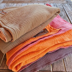 Pu&ograve; includere: Un set di quattro coperte in mussola di cotone in tonalit&agrave; di marrone, arancione e rosa. Le coperte sono piegate e disposte su una superficie in legno.