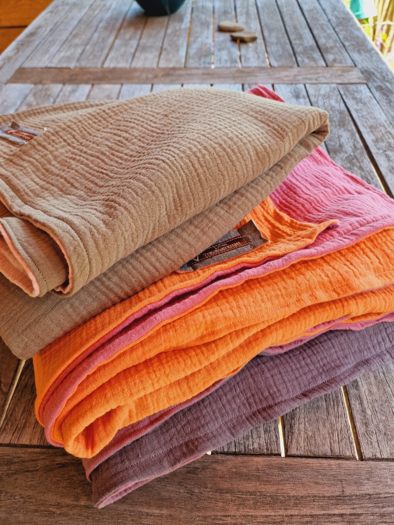 Pu&ograve; includere: Un set di quattro coperte in mussola di cotone in tonalit&agrave; di marrone, arancione e rosa. Le coperte sono piegate e disposte su una superficie in legno.