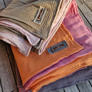 Pu&ograve; includere: Un mucchio di quattro coperte in mussola per bambini in diversi colori, tra cui arancione, rosa e grigio. Le coperte sono piegate e hanno un'etichetta che dice "Liebe Tr&auml;umchen".