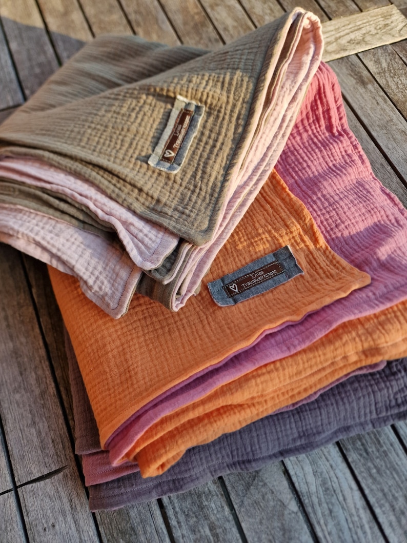 Pu&ograve; includere: Un mucchio di quattro coperte in mussola per bambini in diversi colori, tra cui arancione, rosa e grigio. Le coperte sono piegate e hanno un'etichetta che dice "Liebe Tr&auml;umchen".