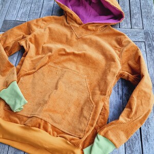May include: A long-sleeve hooded sweatshirt made of orange corduroy fabric. The hood features a bright purple lining. The garment includes a large front kangaroo pocket, a solid orange hem, and light green ribbed cuffs at the end of each sleeve.