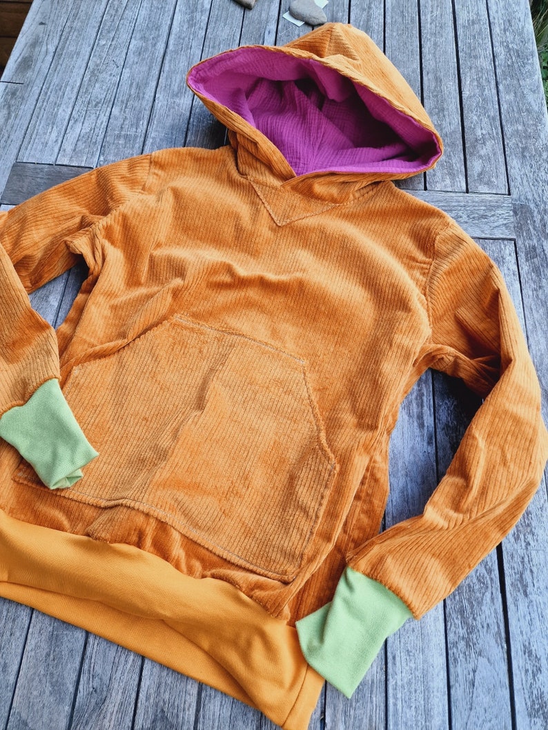 May include: A long-sleeve hooded sweatshirt made of orange corduroy fabric. The hood features a bright purple lining. The garment includes a large front kangaroo pocket, a solid orange hem, and light green ribbed cuffs at the end of each sleeve.