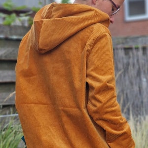 May include: A mustard yellow corduroy hooded sweatshirt featuring a textured ribbed fabric, long sleeves, and a solid mustard yellow hem. The garment includes a hood and is worn over a light green undershirt and blue denim jeans.