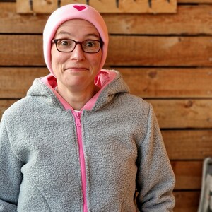 May include: A woman wearing a gray fleece jacket with a pink zipper and pink trim. She is also wearing a pink beanie with a small pink heart on the front.