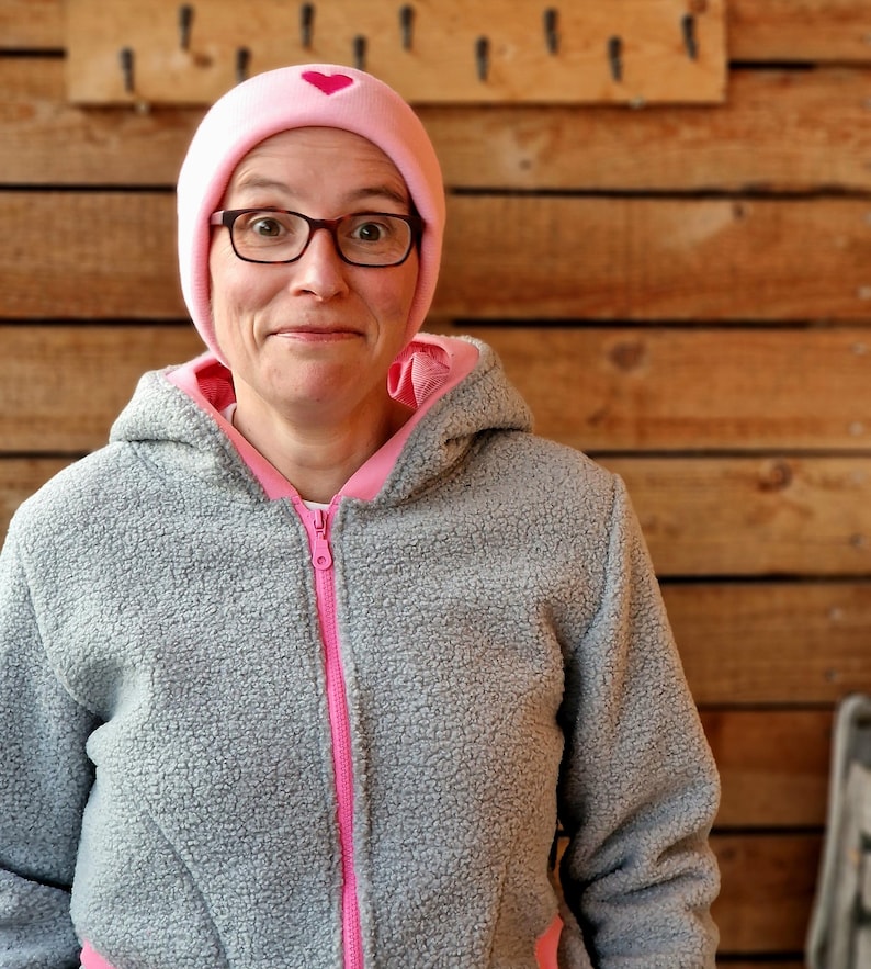 May include: A woman wearing a gray fleece jacket with a pink zipper and pink trim. She is also wearing a pink beanie with a small pink heart on the front.