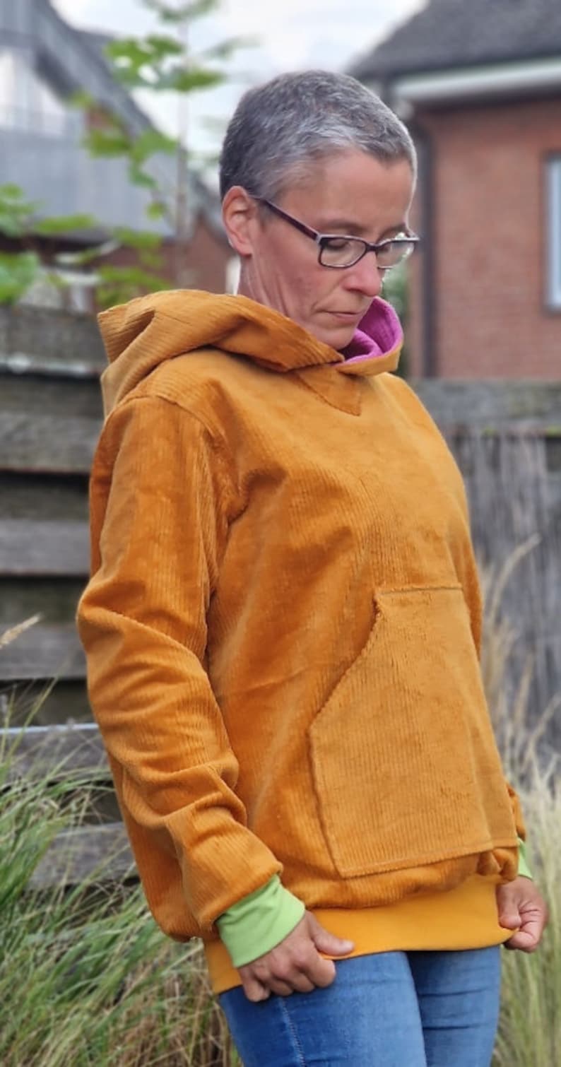 May include: A mustard yellow corduroy hoodie featuring a large front kangaroo pocket, a vibrant pink inner hood lining, and light green ribbed cuffs. The long-sleeved pullover garment has a thick textured fabric finish and a wide hem band.
