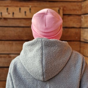 May include: A person wearing a light pink beanie and a gray hooded jacket. The beanie is a solid color, and the jacket has a textured, fuzzy appearance. The background is a wooden wall.