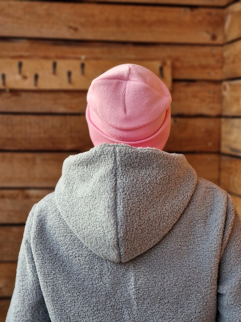 May include: A person wearing a light pink beanie and a gray hooded jacket. The beanie is a solid color, and the jacket has a textured, fuzzy appearance. The background is a wooden wall.