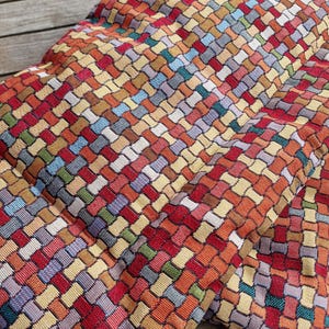 Può includere: Primo piano di un tessuto intrecciato con un motivo a trama di cestino colorato. Il tessuto presenta un mix di tonalità rosse, arancioni, gialle, verdi, blu e viola, creando una superficie vibrante e strutturata. Il tessuto sembra essere un cuscino.