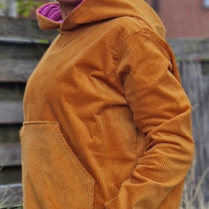 May include: A mustard yellow corduroy hooded sweatshirt featuring a large front kangaroo pocket, a bright pink inner hood lining, lime green sleeve cuffs, a wide mustard yellow hem, and a small teal patch pocket on the lower side.