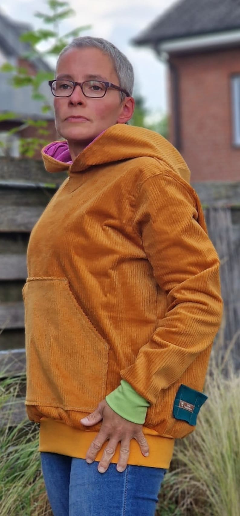 May include: A mustard yellow corduroy hooded sweatshirt featuring a large front kangaroo pocket, a bright pink inner hood lining, lime green sleeve cuffs, a wide mustard yellow hem, and a small teal patch pocket on the lower side.