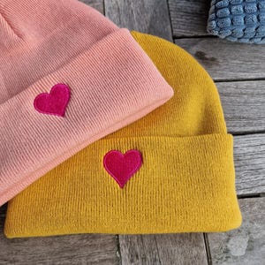 May include: Two knit beanies with heart embroidery. The beanie on top is pink with a pink heart. The beanie on the bottom is yellow with a red heart.