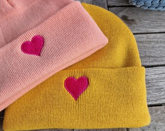 May include: Two knitted beanies with heart embroidery. The beanie on top is pink with a pink heart, and the beanie on the bottom is yellow with a red heart.