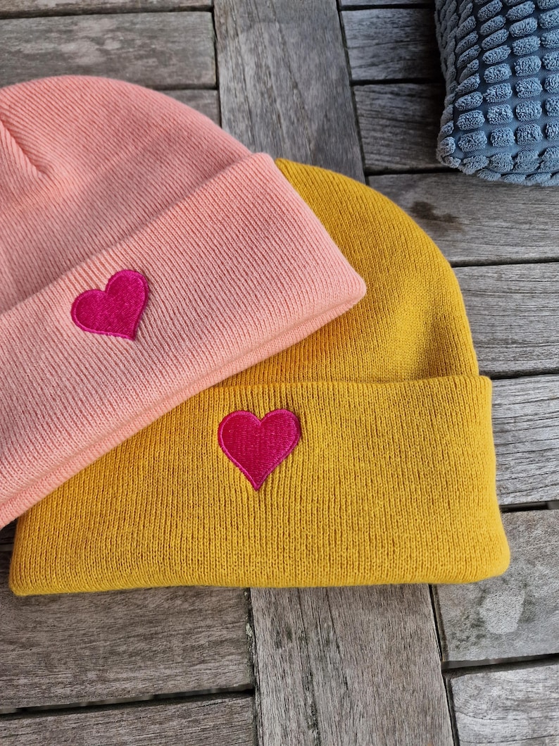 May include: Two knit beanies with heart embroidery. The beanie on top is pink with a pink heart. The beanie on the bottom is yellow with a red heart.