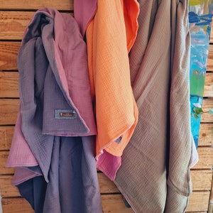 Pu&ograve; includere: Tre morbide coperte per bambini in tessuto testurizzato, in diversi colori: viola, arancione e marrone. Le coperte sono piegate e appese a una parete di legno.