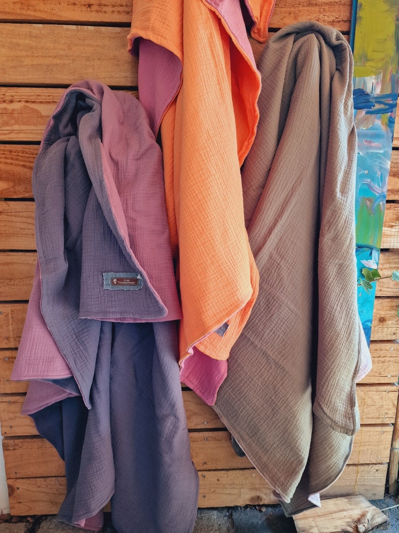 Pu&ograve; includere: Tre morbide coperte per bambini in tessuto testurizzato, in diversi colori: viola, arancione e marrone. Le coperte sono piegate e appese a una parete di legno.