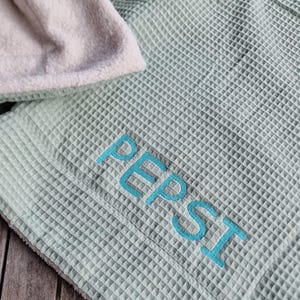 Könnte beinhalten: Eine hellgrüne Waffeldecke mit dem Wort "PEPSI" in Blau gestickt. Die Decke hat ein weiches, cremefarbenes Futter und liegt auf einer Holzoberfläche.