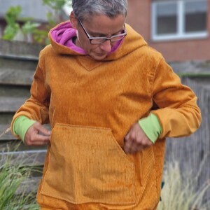 May include: A mustard yellow corduroy hooded sweatshirt featuring a large front kangaroo pocket, a bright pink inner hood lining, and lime green ribbed cuffs. The garment has a wide mustard yellow hem band and long sleeves.