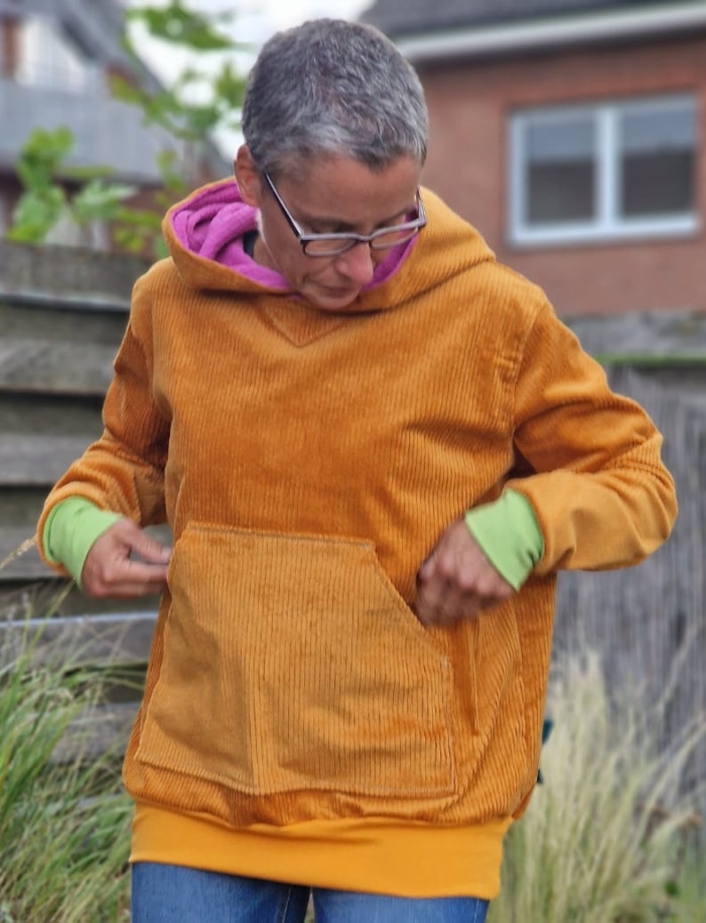 May include: A mustard yellow corduroy hooded sweatshirt featuring a large front kangaroo pocket, a bright pink inner hood lining, and lime green ribbed cuffs. The garment has a wide mustard yellow hem band and long sleeves.