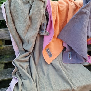Pu&ograve; includere: Tre coperte di mussola di cotone piegate e impilate su una panca di legno. La coperta superiore &egrave; di colore verde chiaro, la coperta centrale &egrave; di colore arancione chiaro e la coperta inferiore &egrave; di colore viola chiaro. Ogni coperta ha un piccolo cartellino con la parola "Muslin" stampata su di essa.