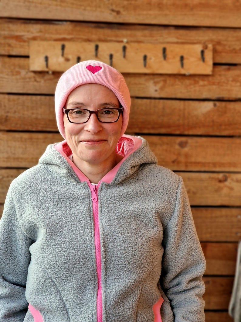 May include: A woman wearing a gray fleece jacket with pink trim and a pink beanie with a pink heart on it. She is wearing glasses and smiling.