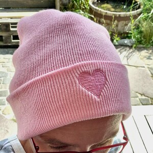 May include: A light pink beanie hat with a folded brim. The hat features a stitched heart design on the front. The hat is made of a soft, ribbed knit material. The beanie is a casual accessory.