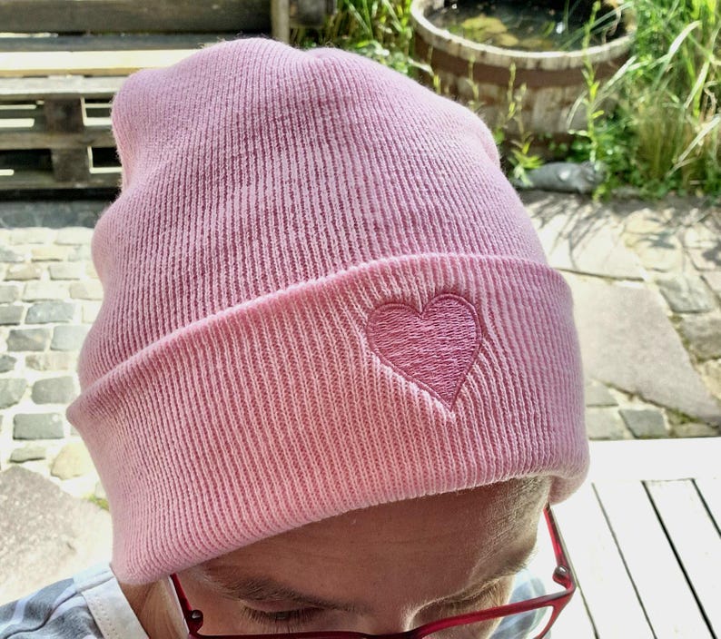 May include: A light pink beanie hat with a folded brim. The hat features a stitched heart design on the front. The hat is made of a soft, ribbed knit material. The beanie is a casual accessory.