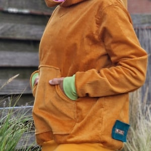 May include: A mustard yellow corduroy hoodie featuring a large front kangaroo pocket, a hood with a bright pink inner lining, lime green sleeve cuffs, and a solid yellow hem. A small rectangular teal fabric tag is attached to the lower side.