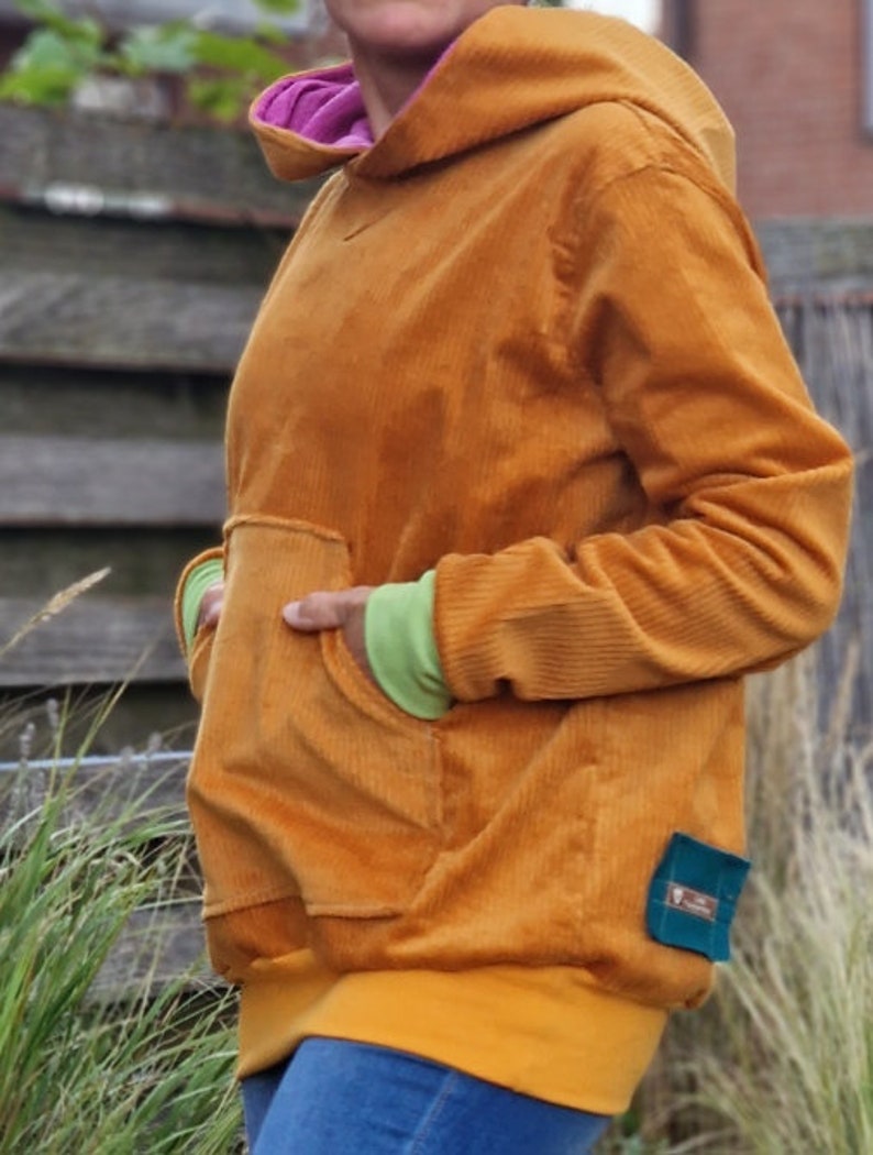 May include: A mustard yellow corduroy hoodie featuring a large front kangaroo pocket, a hood with a bright pink inner lining, lime green sleeve cuffs, and a solid yellow hem. A small rectangular teal fabric tag is attached to the lower side.