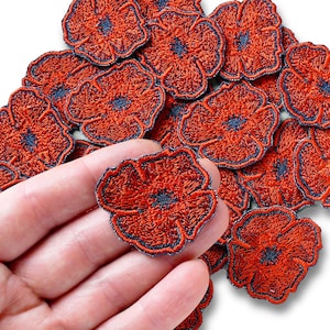Könnte beinhalten: Eine Sammlung gestickter Mohnblumen-Patches. Die Patches sind hauptsächlich rot mit einem dunkelblauen Zentrum und Rand. Die Patches haben die Form einer Mohnblume.