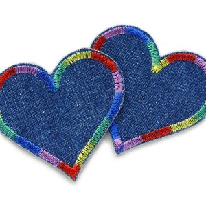 Könnte beinhalten: Zwei herzförmige Aufnäher aus Denim in Dunkelblau mit regenbogenfarbigen, gestickten Rändern. Die Aufnäher überlappen sich leicht und erzeugen einen geschichteten Effekt. Die Regenbogenfarben setzen einen lebendigen Kontrast.
