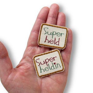 Könnte beinhalten: Zwei rechteckige bestickte Aufnäher mit cremefarbenem Stoffuntergrund und goldfarbenen Stickrändern. Ein Aufnäher zeigt den grünen und roten Schriftzug "Super held", der andere den grünen und roten Text "Super heldin" mit einem kleinen roten Herz.