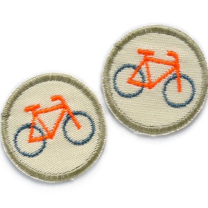 May include: Two round embroidered iron-on patches with a white background and a grey border. Each patch features an orange bicycle with blue wheels.