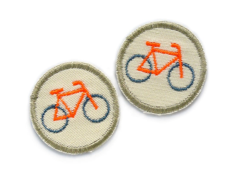 May include: Two round embroidered iron-on patches with a white background and a grey border. Each patch features an orange bicycle with blue wheels.