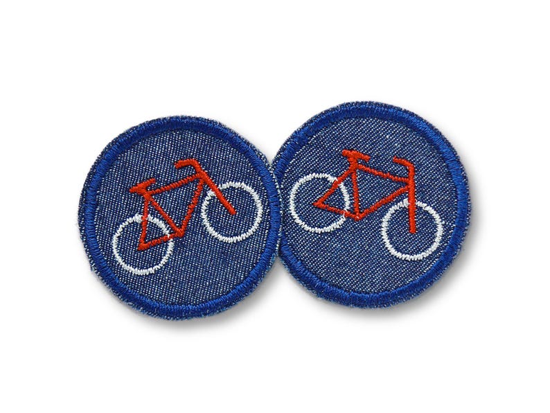 May include: Two round denim patches with a red bicycle design and white wheels. The patches have a blue border and are set against a white background. These patches are ideal for adding to clothing or accessories.
