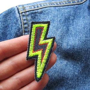 Könnte beinhalten: Ein gestickter Blitz-Patch in Gelb und Rot. Der Patch befindet sich auf einem Jeans-Hintergrund.