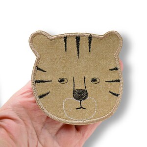 Könnte beinhalten: Gestickter Aufnäher in Form eines Tigergesichts. Der Aufnäher ist beige mit schwarzen Streifen und Gesichtsdetails. Nase und Mund des Tigers sind mit weißem Faden umrandet. Für Kleidung oder Accessoires.