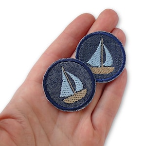 Könnte beinhalten: Zwei blaue Jeansstoff-Patches mit gestickten weißen Segelbooten und braunen Rümpfen.
