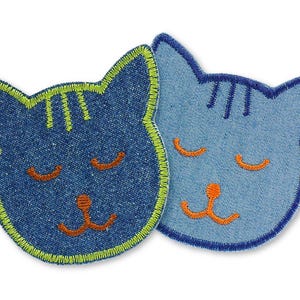 Könnte beinhalten: Zwei blaue Jeans-Katzenkopf-Patches mit gestickten Details. Eine Katze hat grüne Nähte an den Rändern und die andere blaue Nähte.