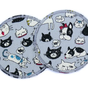 Könnte beinhalten: Zwei runde, graue Stoff-Stillpads mit einem schwarz-weißen Katzen- und Hundemuster. Die Pads haben einen weißen Rand mit einem schwarzen, gesteppten Rand.