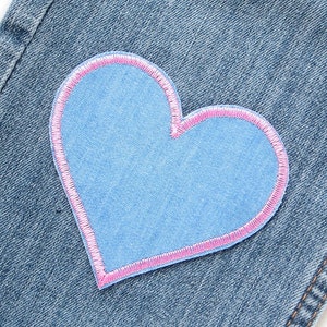 Pu&ograve; includere: Primo piano di una toppa blu a forma di cuore con un bordo cucito rosa, cucita su tessuto denim. La toppa &egrave; di una tonalit&agrave; di blu pi&ugrave; chiara rispetto al denim. Il denim presenta cuciture visibili.