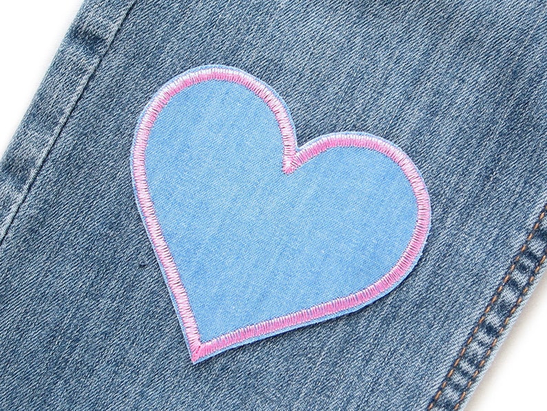 Pu&ograve; includere: Primo piano di una toppa blu a forma di cuore con un bordo cucito rosa, cucita su tessuto denim. La toppa &egrave; di una tonalit&agrave; di blu pi&ugrave; chiara rispetto al denim. Il denim presenta cuciture visibili.